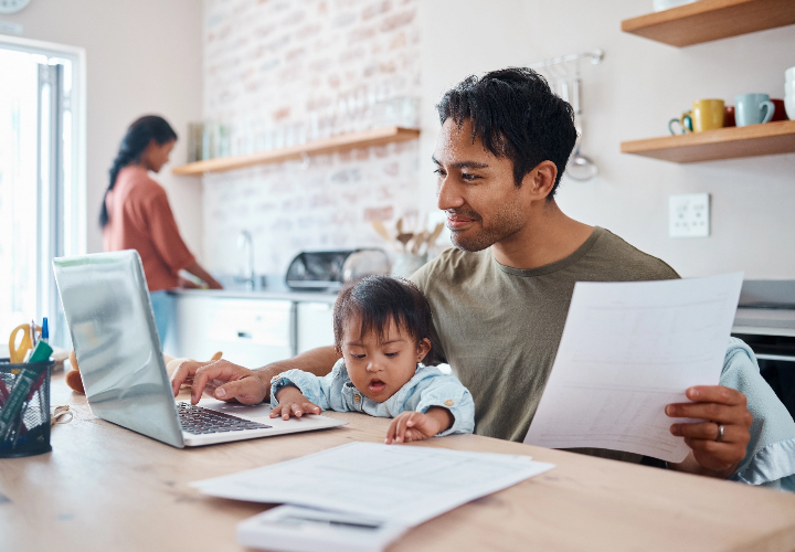 Father working on laptop with son on his lap, sitting at the kitchen table.